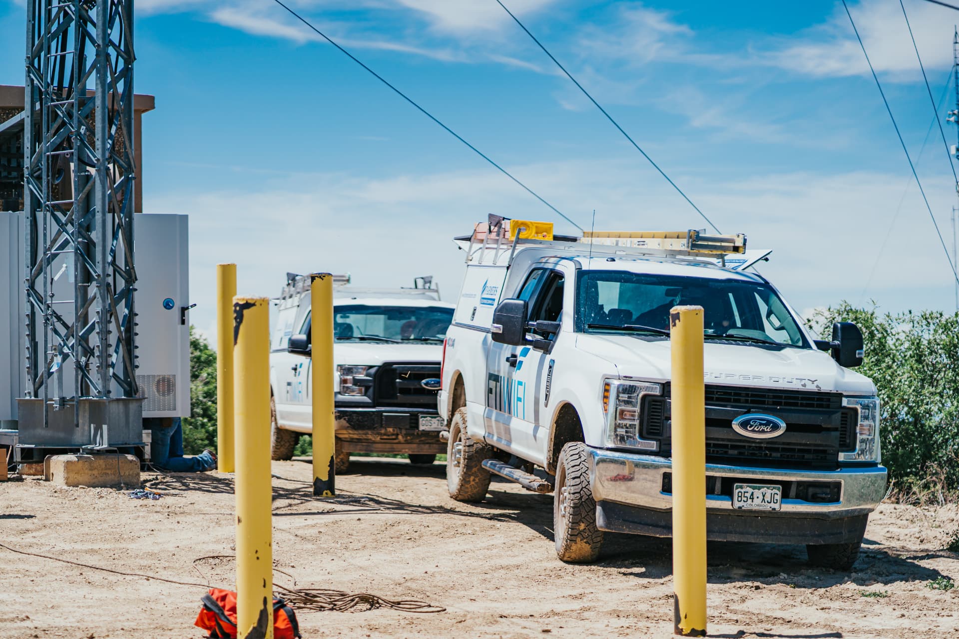 Two FTI WIFI service trucks parked at a communications tower site under a blue sky